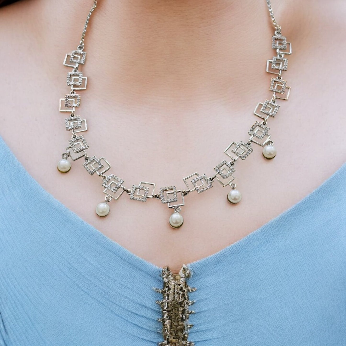 Necklace with pearls and square designs on a person wearing a blue top.