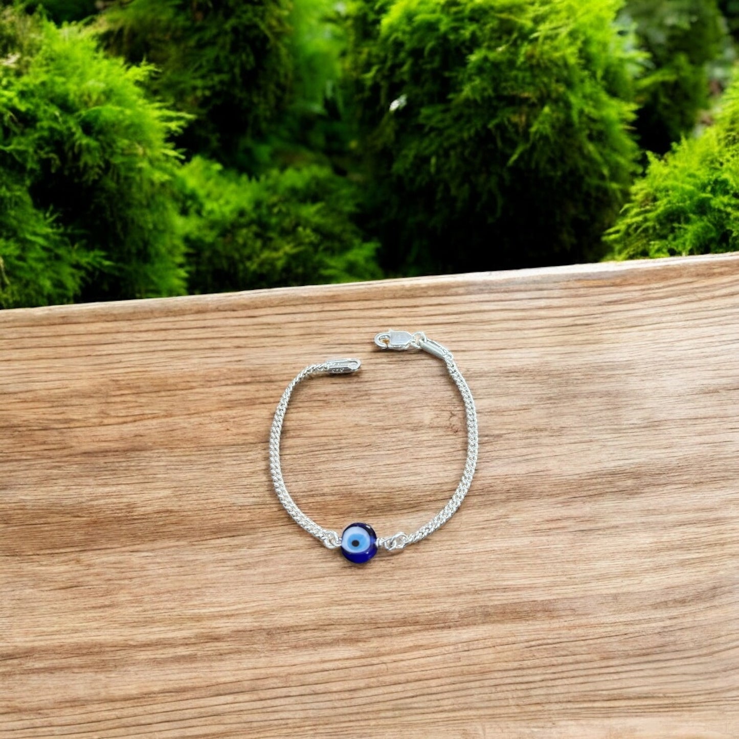 Silver bracelet with a blue eye charm on a wooden surface with greenery in the background