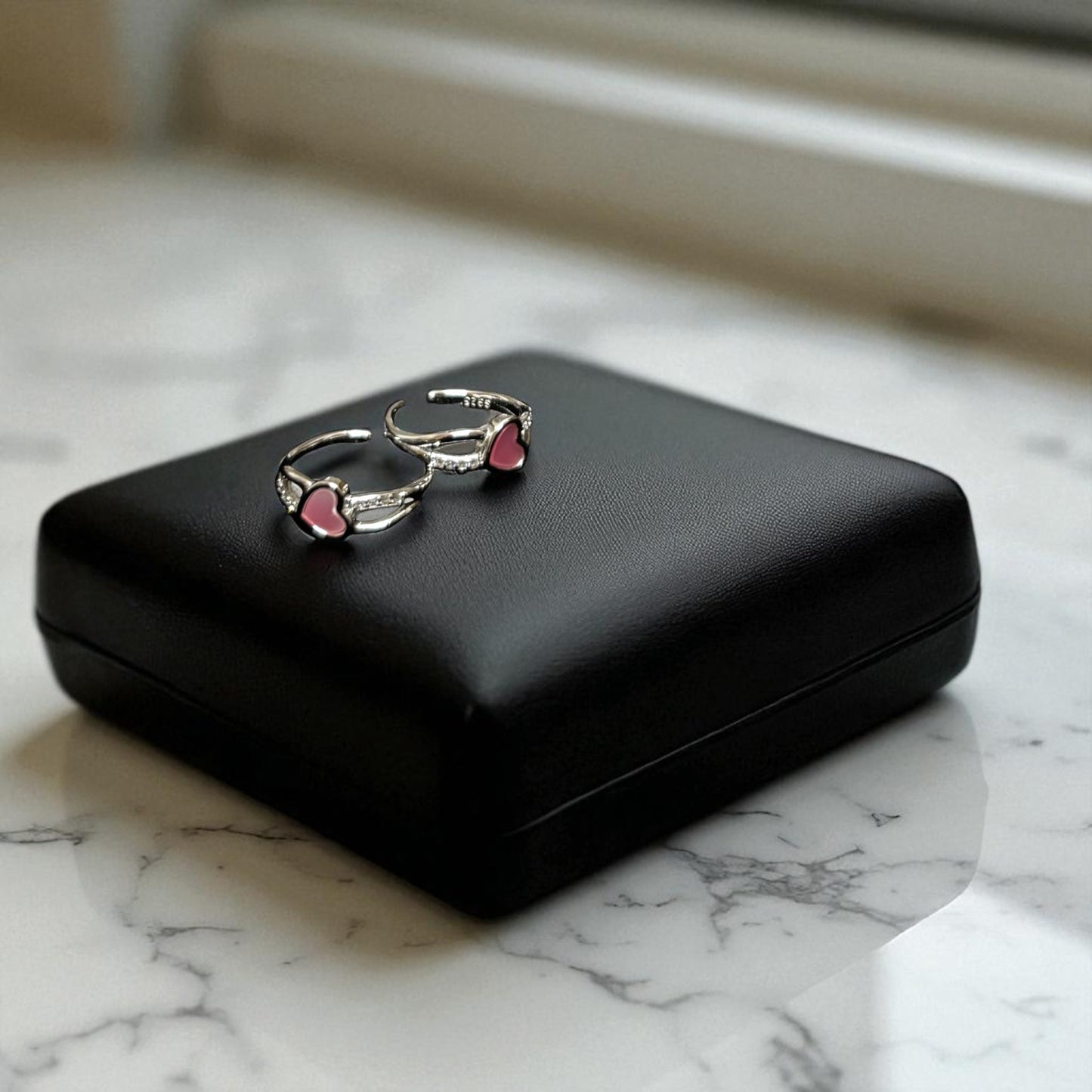 Pair of silver earrings with pink heart details on a black case against a marble background