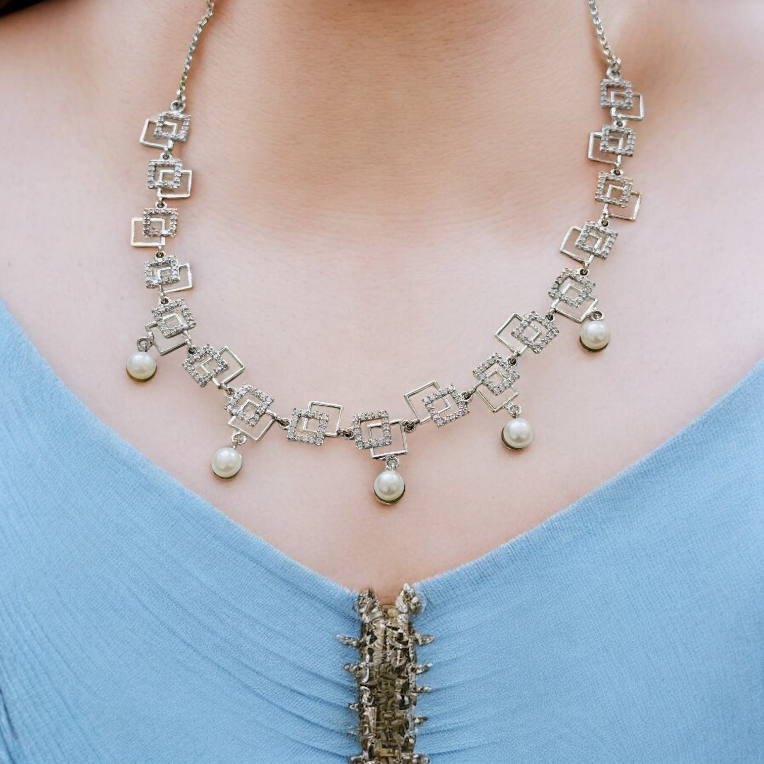 Necklace with pearls and square designs on a person wearing a blue top.
