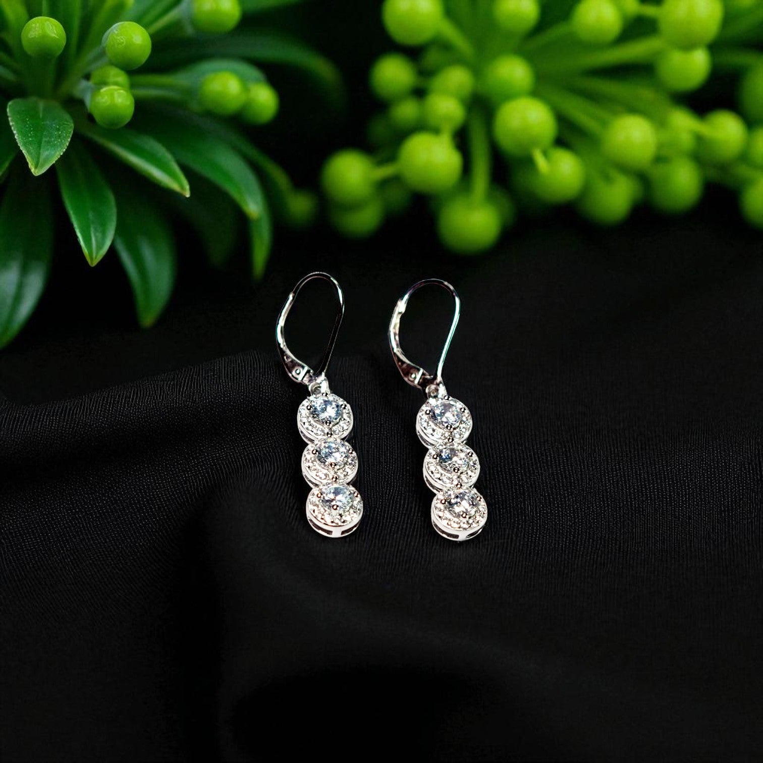 Silver earrings with clear stones on a black surface with green foliage in the background